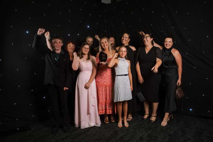 A group of award recipients dressed in formal attire pose together in front of a black star‑themed backdrop, holding a trophy for the 2025 sports awards.